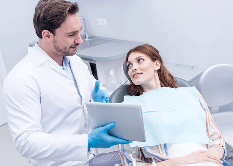 A male dentist wearing blue gloves discusses treatment options with a smiling female patient in a dental clinic, using a tablet to explain the procedure, highlighting dental tourism and patient care.