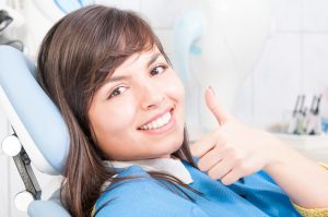 A happy female patient sitting in a dental chair, smiling and giving a thumbs-up, indicating a positive dental experience