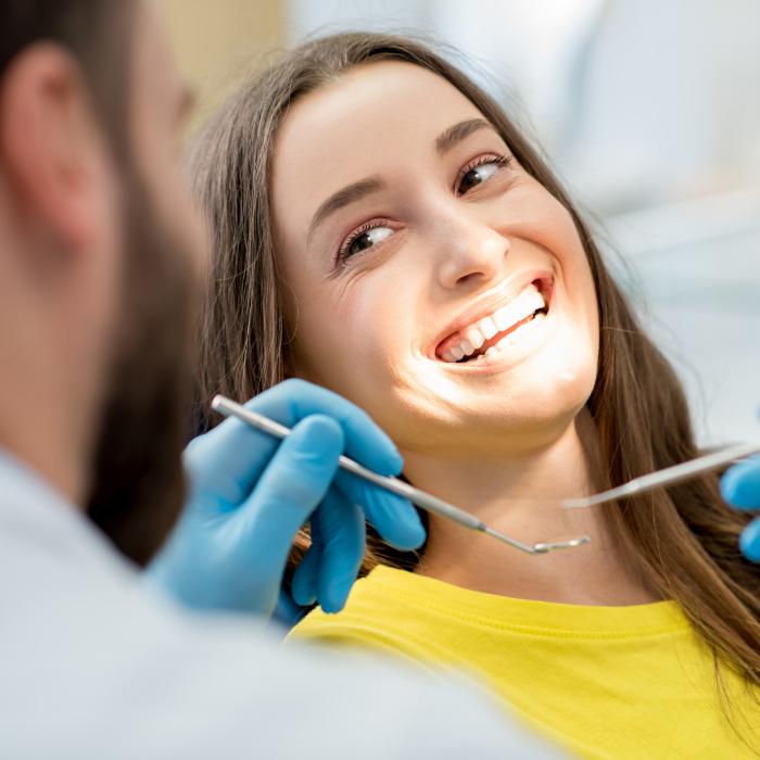A happy young woman in a yellow shirt smiling brightly during a dental check-up. The dentist, wearing blue gloves, holds dental tools while examining her teeth, highlighting a positive dental care experience.