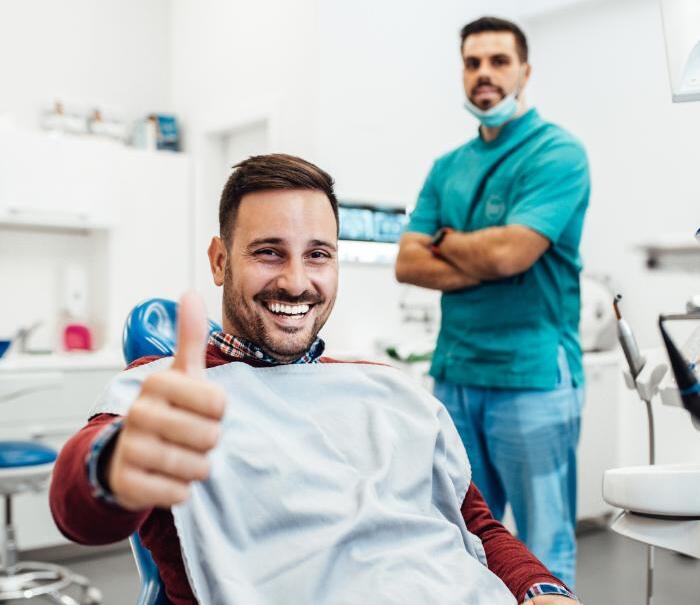 A happy male patient sitting in a dental chair giving a thumbs-up after treatment, while a dentist in scrubs stands confidently in the background. The image highlights a successful and positive dental care experience.