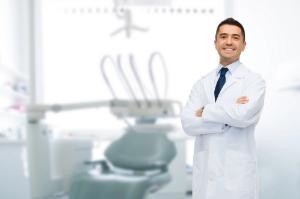 A smiling male dentist in a white coat stands confidently with crossed arms in a modern dental clinic, with a dental chair and equipment in the background.
