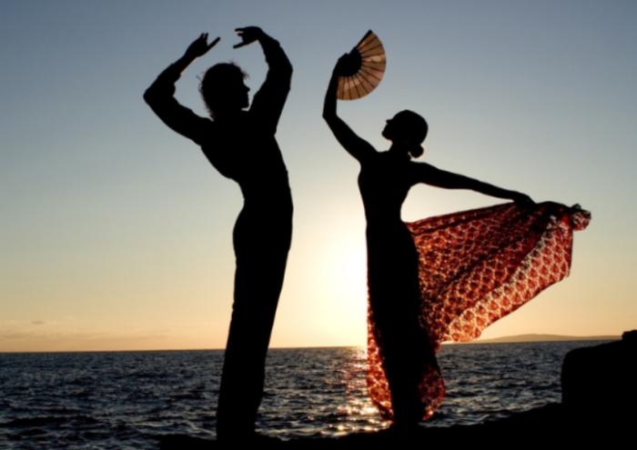 Silhouettes of two flamenco dancers performing against a sunset by the sea. One dancer gracefully holds a fan, while the other strikes an elegant pose, capturing the essence of Spanish culture and tradition.