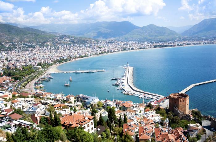 A scenic aerial view of Alanya, Turkey, showcasing the Red Tower, marina, and coastline, with the city nestled between the Mediterranean Sea and lush mountains.