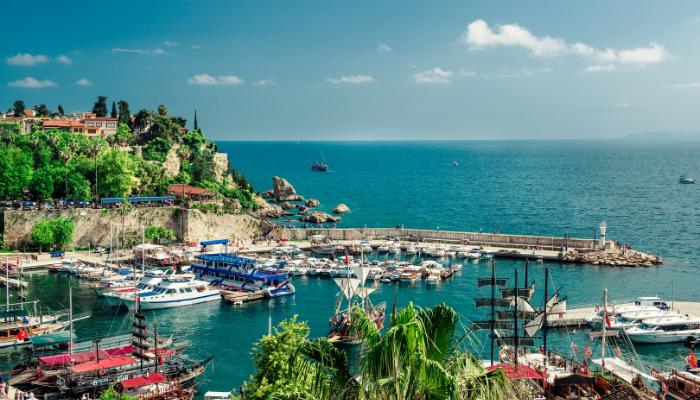 A picturesque view of Antalya's Old Harbor in Turkey, featuring boats docked along the turquoise Mediterranean waters, surrounded by lush greenery and historic cliffs under a bright blue sky.