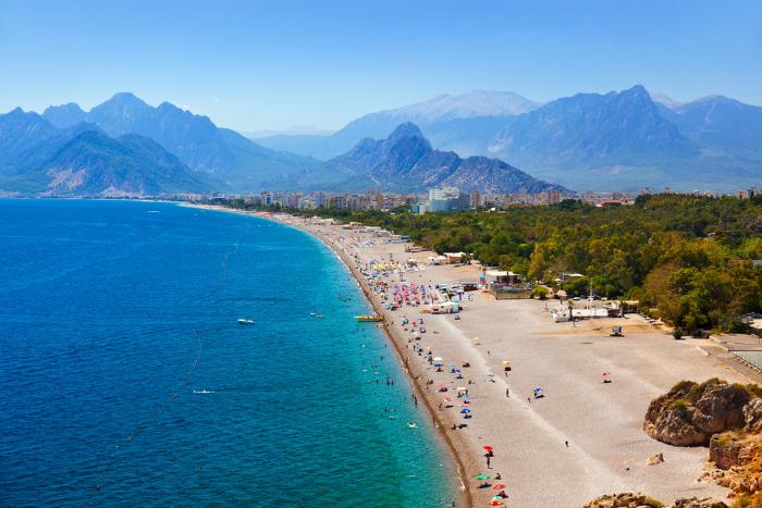 A stunning aerial view of Konyaaltı Beach in Antalya, Turkey, with turquoise waters, a long pebble shoreline, and the Taurus Mountains in the background. Tourists are sunbathing and enjoying the beach, while the cityscape of Antalya stretches along the coast.