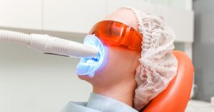 A patient undergoing professional teeth whitening treatment with a blue LED light device in a dental clinic. The patient is wearing protective orange-tinted glasses and a hair cap for safety during the procedure.