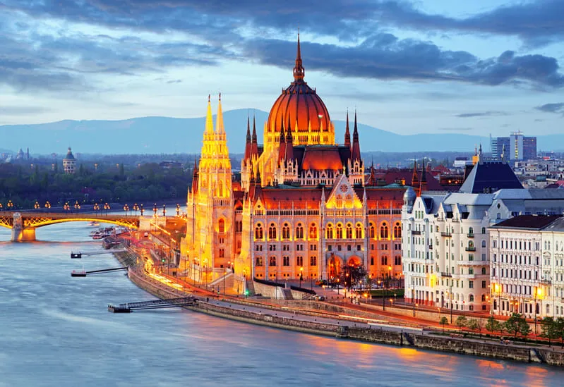 A breathtaking evening view of the Hungarian Parliament Building in Budapest, Hungary, illuminated along the banks of the Danube River. The iconic neo-Gothic architecture stands out against a dramatic sky, with the Margaret Bridge and city lights enhancing the scenic backdrop.
