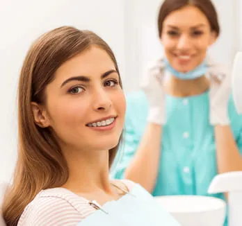 A happy female patient with a bright smile sitting in a dental chair after treatment, with a smiling dentist in the background, highlighting a positive dental care experience.