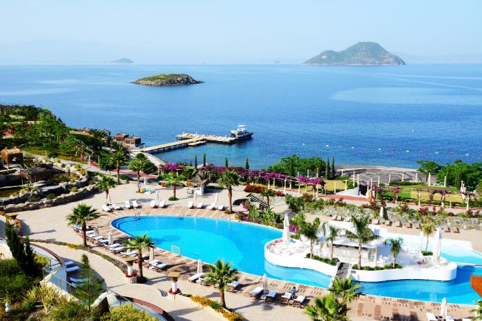 A luxury seaside resort in Bodrum, Turkey, featuring a large swimming pool, palm trees, and a scenic view of the Aegean Sea with small islands in the background.