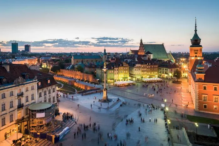 A scenic evening view of Castle Square in Warsaw, Poland, featuring the Sigismund's Column in the foreground, historic buildings, and a lively crowd. The Royal Castle and old town architecture are illuminated, creating a vibrant atmosphere.