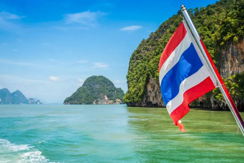 A scenic view of the turquoise waters and lush green limestone cliffs of Thailand, with a Thai flag waving on a boat in the foreground under a bright blue sky.