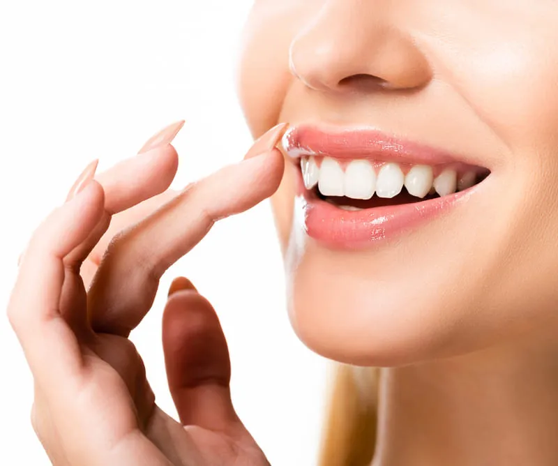 A close-up of a woman with a bright, white smile touching her lips, highlighting smooth skin and perfect teeth, possibly enhanced by veneers or teeth whitening.